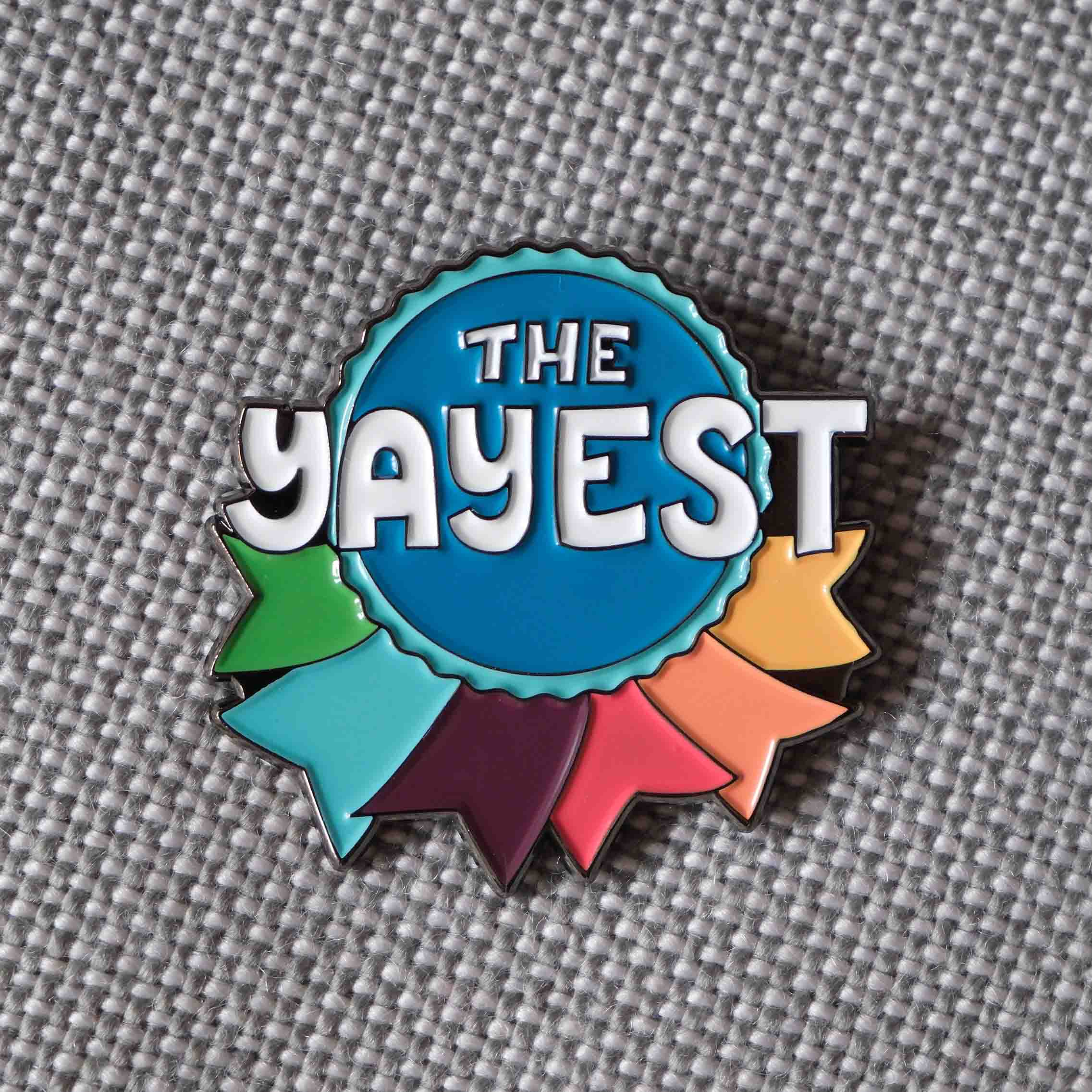 An enamel pin with a rainbow ribbon design that says "The Yayest"