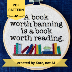 PDF pattern created by Kate, not AI. A book worth banning is a book worth reading. Quote hand embroidered in black serif font. Below the text is a bookshelf with several different colored books, as well as a light blue planter holding a light green plant. Sewn on white cotton. Framed in a light wood embroidery hoop.