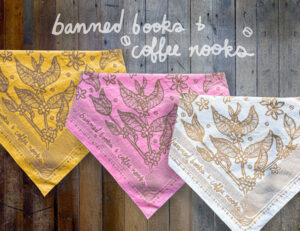 Sugar Camp Banned Books & Coffee Nooks Bandana in flower power pink, groovy gold, and cream, folded and overlapping on weathered wood floor.