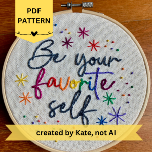 PDF Pattern created by Kate, not AI. Be Your Favorite Self is embroidered in a script font on cream fabric with gold threads woven in. Favorite is sewn in rainbow colors, and stars surround the quote. This wall art is framed in a light wood embroidery frame.