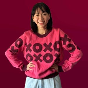 A woman wearing a pink sweater with a dark red XOXO design that goes across the front.