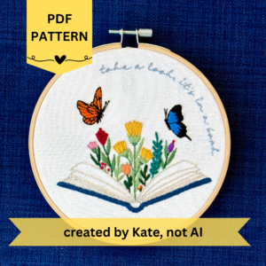 PDF pattern created by Kate, not AI. Photo of a hand embroidered design inspired by Reading Rainbow. There is an open book with a rainbow of flowers growing from it. Hovering above are an orange and black butterfly and a bright blue and black butterfly. The top right curve of the pattern reads, "take a look, it's in a book," sewn in light blue embroidery thread. Displayed in a light wooden embroidery hoop suitable for wall art. Fabric is white cotton with white flowers printed on it.
