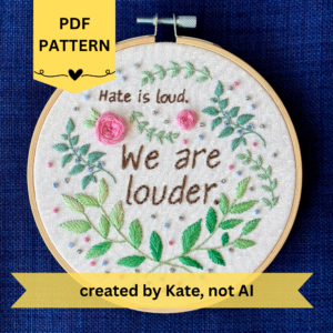 PDF pattern created by Kate, not AI. Hate is loud. We are louder. Quote from Somewhere Beyond the Sea by TJ Klune. Text hand embroidered in chocolate brown. Surrounded by green leaves & vines, with pink roses, blue berries, & metallic silver dots. Framed in light wood embroidery hoop
